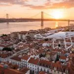Drone branco a voar sobre os telhados de Lisboa com a Ponte 25 de Abril e o Tejo ao fundo, ao pôr do sol